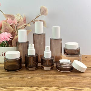 Op de afbeelding: Een verzameling doorschijnende bruine glazen cosmetische verpakkingen met witte deksels en pompdoseerders. De set bevat twee hoge cilindrische flessen, twee kleinere pompflessen en drie ronde potjes van verschillende groottes.