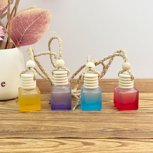 Puede incluir: Cuatro pequeñas botellas de vidrio cuadradas en amarillo, morado, azul y rojo, cada una con una tapa de madera y una cuerda para colgar. Las botellas están dispuestas sobre una superficie de madera, lo que sugiere un producto decorativo o aromático.