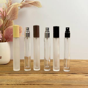 Puede incluir: Seis frascos de perfume de vidrio transparente con atomizadores y varios estilos de tapones, incluyendo madera y negro. Los frascos están dispuestos sobre una superficie de madera, con un fondo blanco y acentos florales.