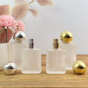Peut inclure: Deux flacons de parfum rectangulaires en verre dépoli avec embouts vaporisateurs. Un flacon possède un bouchon sphérique argenté et l'autre un bouchon sphérique doré. Deux bouchons sphériques assortis, un argenté et un doré, sont posés devant.