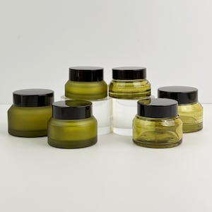 Puede incluir: Seis tarros de vidrio verde oliva con tapas negras. Los tarros son de varios tamaños y formas, algunos con un acabado esmerilado. Los tarros están dispuestos sobre una superficie blanca, lo que sugiere un producto para el cuidado de la piel o cosmético.