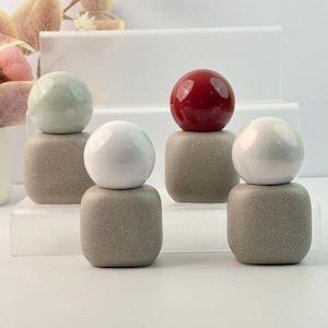 Puede incluir: Cuatro objetos decorativos. Cada uno tiene una base cuadrada y moteada y una parte superior esférica. Las partes superiores son en tonos de blanco, verde y rojo. Los objetos están dispuestos sobre una superficie blanca con un fondo neutro.