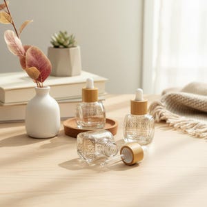 May include: Clear glass dropper bottles with bamboo lids are arranged on a light wood surface. One bottle is lying on its side with the dropper exposed. A small white vase with a sprig of leaves and a succulent plant in a concrete pot are in the background.