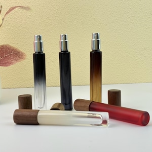 Puede incluir: Una colección de frascos de perfume o aceites esenciales en varios colores. Los frascos tienen atomizadores plateados y tapas de madera. Los colores incluyen transparente, negro y un degradado de negro a marrón. Un frasco es rojo.