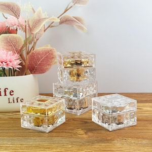 May include: A collection of square, clear acrylic storage boxes with faceted, diamond-like textures on the sides. Three boxes are stacked vertically, while two individual boxes sit beside them. The lids feature metallic gold and silver accents.