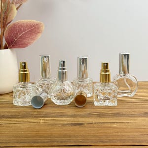 May include: A collection of clear glass perfume bottles with silver and gold spray tops. The bottles have various shapes, including round, square, and heart-shaped designs. Some bottles have their caps removed, revealing the spray mechanisms.