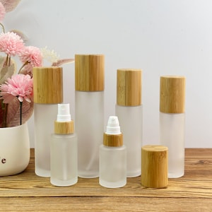 May include: A collection of frosted glass bottles and jars featuring light brown bamboo lids. The set includes various sizes of cylindrical containers, some with white pump dispensers and others with simple caps, arranged in a group.