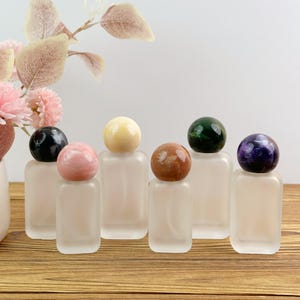 Puede incluir: Seis frascos de perfume rectangulares de vidrio esmerilado con tapas esféricas de piedra en negro, rosa, crema, marrón, verde oscuro y morado. Los frascos están dispuestos en fila, cada uno con un cuerpo translúcido liso y un remate de piedra pulida.