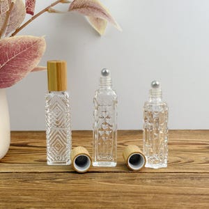 Op de afbeelding: Drie doorzichtige glazen rollerflessen met diamantpatronen. Eén heeft een bamboe dop, twee hebben zilveren rollerball toppen. De flessen staan op een houten oppervlak, met een witte achtergrond en decoratieve plant.