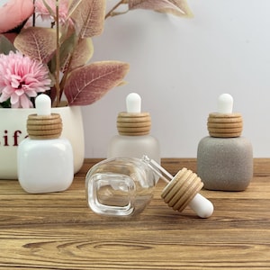 Puede incluir: Una colección de frascos cuentagotas cuadrados de vidrio con tapones de madera y blancos. Una botella es transparente, otras son blancas y grises. Las botellas se exhiben sobre una superficie de madera, con un jarrón de flores rosas en el fondo.