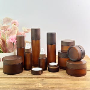 Op de afbeelding: Een collectie cosmetische verpakkingen van amberkleurig glas met donkerbruine houten deksels. De set bevat diverse maten cilindrische flessen met pompdoseerders en ronde potten met schroefdeksels, gerangschikt op een houten ondergrond.