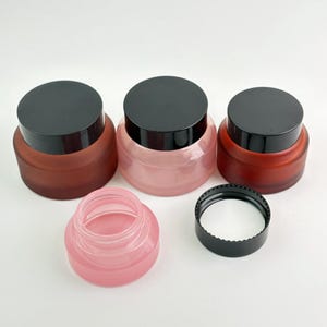 May include: A collection of cosmetic jars in various shades of pink and brown, with black lids. One jar is open, revealing the interior. The jars have a frosted appearance, and the lids are solid black.