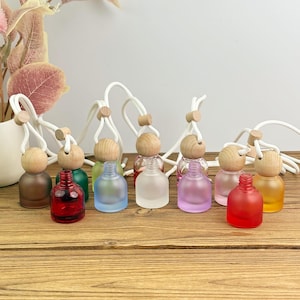 May include: A collection of colorful, small glass bottles with wooden tops and white cords for hanging. The bottles are in various colors, including brown, red, green, blue, clear, purple, pink, and yellow. They are arranged on a wooden surface.