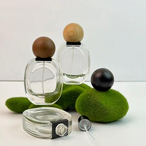 Three transparent glass perfume bottles with wooden spherical caps are present: one stands upright at the front left of the scene; another rests slightly higher to its right rear, both resting on moss-covered stones. A third transparent perfume bottle without its cap lies on its side at the lower left of the scene, revealing its opening and black spray mechanism. Its black spherical cap rests atop a green stone ornament nearby.