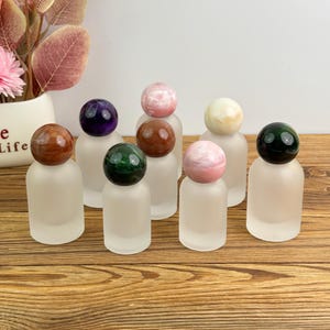 May include: A collection of frosted glass perfume bottles, each topped with a different colored gemstone sphere. The gemstones include brown, purple, green, pink, and cream-colored stones. The bottles are arranged on a wooden surface.