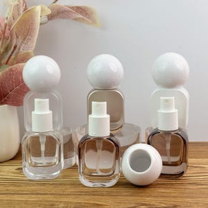 May include: Three square glass spray bottles with rounded corners and white spherical caps. Two bottles are upright with white spray nozzles, while one white spherical cap rests separately in front of the others on a wooden surface.