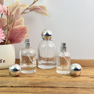 May include: Clear glass perfume bottles with silver sprayers and spherical caps. The bottles are arranged on a wooden surface, with a vase of pink flowers and foliage in the background. Two silver spheres are also present.