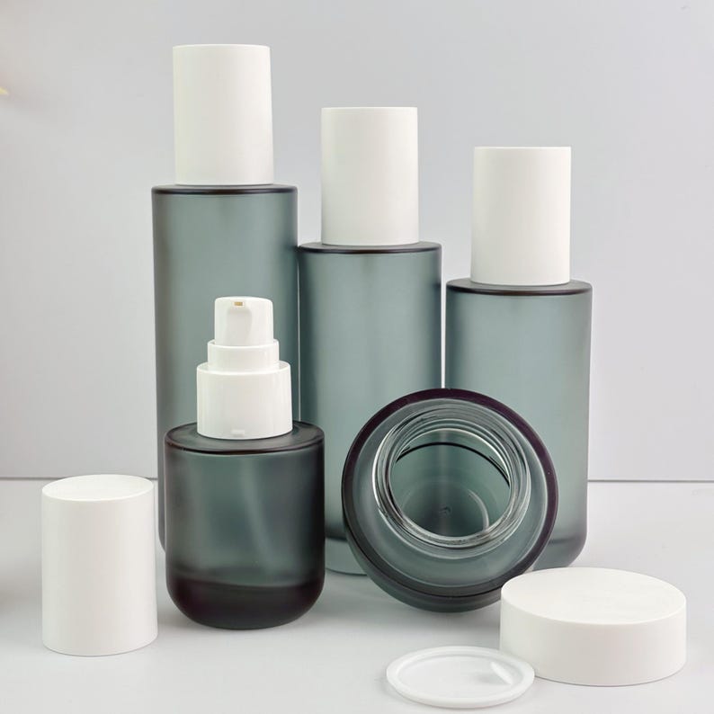 May include: A selection of grey glass cosmetic bottles with white lids and a pump dispenser. The bottles are of various sizes and shapes, with a dark base and a frosted finish. One bottle is open, showing the inside.
