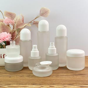Peut inclure: Une collection de contenants cosmétiques en verre dépoli avec des couvercles blancs, comprenant des flacons cylindriques hauts, des flacons à pompe plus petits et des pots ronds. L'ensemble présente diverses tailles et types de fermetures.