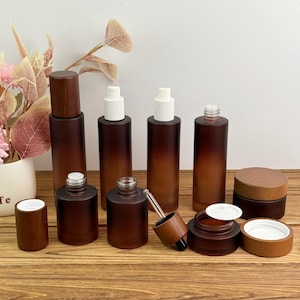 May include: A collection of brown glass cosmetic bottles and jars with wooden lids and white pumps. The bottles vary in size and shape, including a dropper bottle, a small jar, and several cylindrical bottles. The set is arranged on a wooden surface.