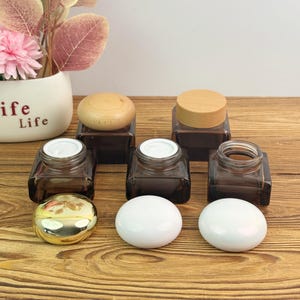 Könnte beinhalten: Quadratische, dunkelbraune Glasbehälter mit Holz- und weißen Deckeln. Einige Gläser sind geöffnet und zeigen eine weiße Creme. Ein goldener und weißer Deckel sowie zwei weiße, ovale Deckel sind ebenfalls vorhanden. Eine Vase mit dem Wort "Life" befindet sich im Hintergrund.