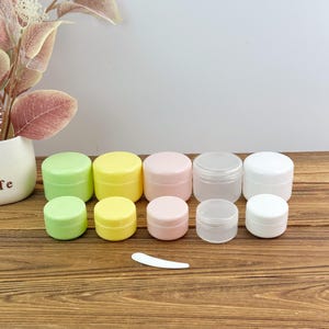 May include: A collection of ten small, round cosmetic jars arranged in two rows, featuring colors of light green, yellow, pale pink, translucent, and white. Each jar has a matching screw-on lid. A single small, white, curved plastic spatula is included.