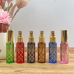 Könnte beinhalten: Sechs zylindrische Parfüm-Sprühflaschen aus Glas mit goldenen Verschlüssen und goldenen Gittermustern. Die Flaschen sind grün, rot, dunkelviolett, bernsteinfarben, blau und rosa und verfügen jeweils über einen feinen Sprühmechanismus.