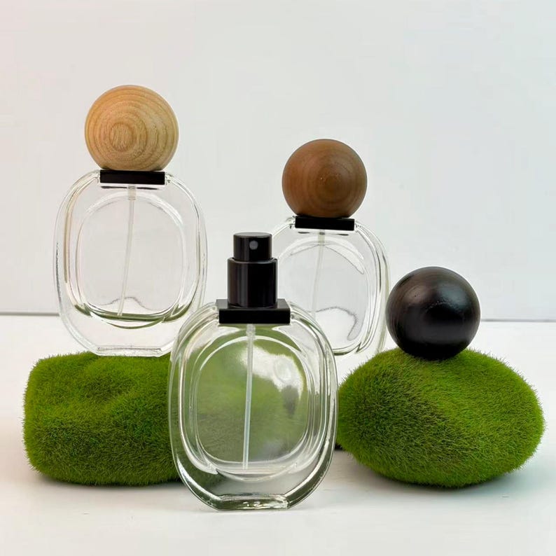 Three identical white transparent glass cosmetic containers, each topped with a differently colored spherical wooden lid—black, brown, and light wood—are arranged in a staggered pattern. Scattered nearby are decorative pieces of green moss.