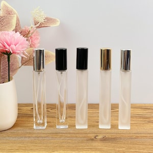 Peut inclure: Cinq flacons de parfum rectangulaires, hauts et fins, alignés. Deux flacons sont en verre transparent avec des bouchons argentés et noirs, tandis que trois sont en verre dépoli avec des bouchons noirs, dorés et argentés. Chaque flacon a un vaporisateur.