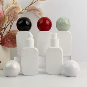 May include: White cosmetic bottles with rounded edges, topped with black, red, and green spherical caps. Two additional white spherical objects are in the foreground. The bottles have spray nozzles. The background is white.