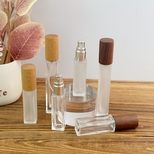 May include: A collection of rectangular glass spray bottles with wooden caps. The set includes frosted and clear glass containers of varying heights, each topped with a silver-toned atomiser pump and a cylindrical wooden lid in light or dark wood tones.