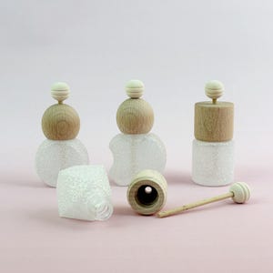May include: A collection of decorative glass bottles with wooden stoppers. The bottles have a clear, textured body with a glittery appearance. The stoppers are made of light-colored wood and topped with a small, round wooden bead. One bottle is lying on its side, with a separate wooden stopper and a small wooden stick.