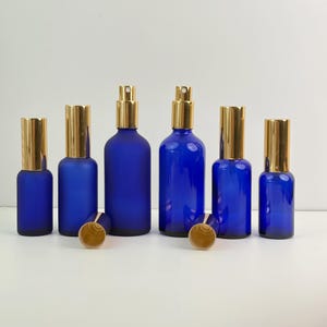 Puede incluir: Una colección de botellas de vidrio azul cobalto con tapas metálicas doradas. Las botellas varían en tamaño y forma, algunas con un acabado esmerilado. Las tapas son cilíndricas y las botellas están dispuestas sobre una superficie blanca.