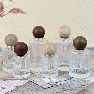 May include: Six clear glass perfume bottles with ribbed sides and wooden ball-shaped stoppers. The stoppers are in various shades of brown and light wood. Each bottle has a gold-colored ring at the base of the stopper.