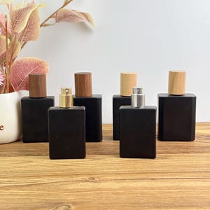 May include: Several black rectangular perfume bottles with wooden and metallic spray tops. The bottles are displayed on a light wooden surface, with a white backdrop and a decorative plant in the corner.