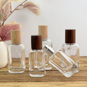 Può includere: Diverse bottiglie di profumo in vetro trasparente con tappi in legno disposte su una superficie di legno. Le bottiglie hanno una forma rettangolare e un vaporizzatore argentato. Alcune hanno tappi in legno chiaro, altre in legno marrone scuro.