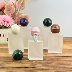 May include: A collection of frosted glass perfume bottles, each topped with a different colored gemstone sphere. The spheres include white, purple, green, pink, and brown. The bottles are arranged on a wooden surface with floral decorations in the background.