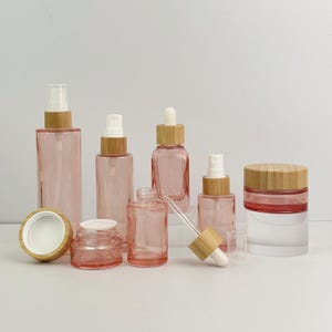 Op de afbeelding: Een verzameling roze glazen cosmeticaflessen en potten met natuurlijke bamboe deksels en sproeiers. De set bevat verschillende maten en vormen, zoals spuitflessen, een druppelfles en een pot met een schroefdeksel.