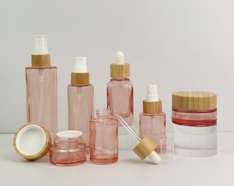 May include: A collection of pink glass cosmetic bottles and jars with natural bamboo lids and sprayers. The set includes various sizes and shapes, such as spray bottles, a dropper bottle, and a jar with a screw-on lid.