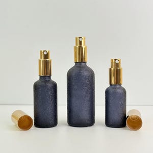 May include: Three dark blue textured glass spray bottles with gold-colored spray tops and matching caps. The bottles are of varying sizes, with one cap detached. The bottles are arranged on a white surface.