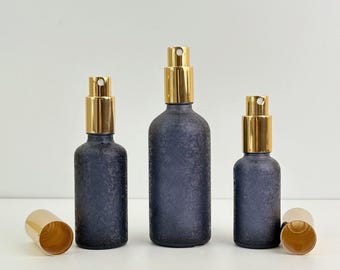 May include: Three dark blue textured glass spray bottles with gold-colored spray tops and matching caps. The bottles are of varying sizes, with one cap detached. The bottles are arranged on a white surface.