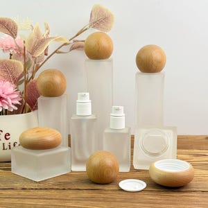 Op de afbeelding: Een verzameling cosmetische flessen en potten van matglas met houten deksels en accenten. De flessen zijn rechthoekig en vierkant, met witte pompknoppen. De houten deksels zijn bolvormig en cilindrisch.
