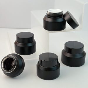 Könnte beinhalten: Mehrere mattschwarze Kosmetikgläser mit passenden Deckeln werden auf einer weißen Oberfläche präsentiert. Ein Glas hat seinen Deckel abgenommen und zeigt einen weißen Innenraum. Die Gläser sind zylindrisch mit abgerundeter Basis, die Deckel flach und kreisförmig.