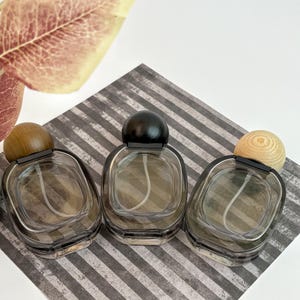 Könnte beinhalten: Drei transparente Parfümflaschen mit abgerundeten, dunkelgrauen Körpern und kugelförmigen Kappen aus Holz oder Schwarz. Die Flaschen sind auf einer grau-weiß gestreiften Oberfläche angeordnet, mit einem Blatt in der oberen linken Ecke. Die Flaschen sind leer.