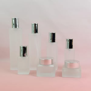 May include: A selection of frosted glass bottles and jars with silver-coloured lids. The containers are in various shapes and sizes, including rectangular and square designs. The background is a soft pink colour.