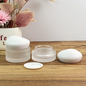 Peut inclure: Deux pots cosmétiques en verre dépoli avec des couvercles blancs et un petit couvercle blanc. Les pots sont vides et prêts à être remplis. Ils sont posés sur une surface en bois.