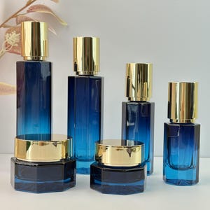 Puede incluir: Un surtido de botellas y frascos de vidrio azul con tapas doradas. Las botellas son rectangulares y cilíndricas, mientras que los frascos son hexagonales. El color azul se gradúa de oscuro a claro. Los recipientes son probablemente para cosméticos o perfumes.