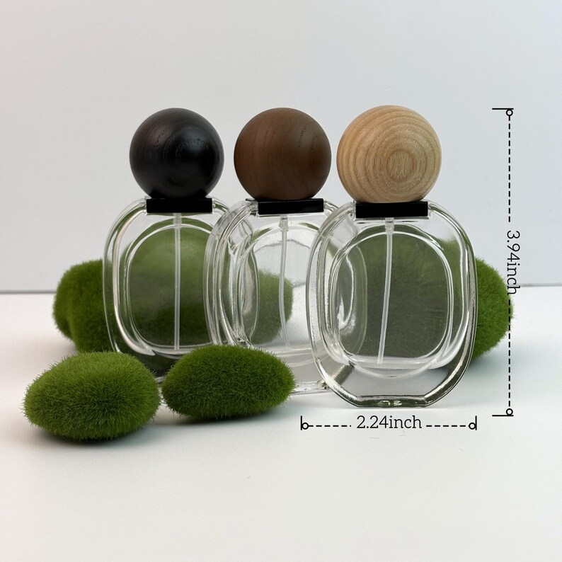 May include: Three clear glass perfume bottles with wooden spherical caps in black, brown, and natural wood tones. The bottles are oval-shaped and measure 3.94 inches high and 2.24 inches wide. Green decorative moss balls are in the foreground.