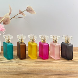 Puede incluir: Un conjunto de seis frascos de perfume rectangulares de vidrio con tapones transparentes tipo cristal y cuellos dorados. Los frascos tienen un acabado esmerilado en colores vibrantes que incluyen verde azulado, marrón, amarillo, magenta, rosa claro y negro mate.