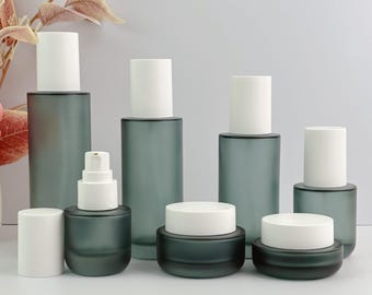 May include: A collection of frosted glass cosmetic containers with white lids. The set includes bottles, jars, and a pump dispenser in a muted teal color, suitable for skincare products.